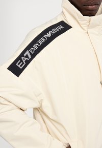 Polstret cremet jakke med høj krave, elastiske manchetter og et sort bånd på skulderen med "EA7 Emporio Armani" logo. Blød tekstur.