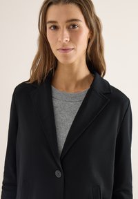 Femme portant un blazer noir sur un pull en maille gris, avec des cheveux bruns raides et une expression neutre, sur un fond clair uni.
