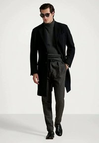 Muž v čiernom kabáte, tmavosivom roláku, plisovaných sivých nohaviciach, čiernych topánkach a slnečných okuliaroch kráča dopredu na bielom pozadí.