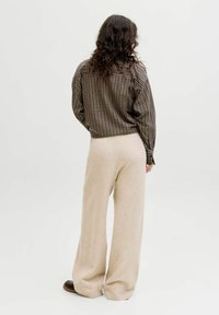 Donna con capelli mossi e scuri che indossa una camicia a scacchi marrone, pantaloni larghi beige e scarpe nere, in piedi di spalle su uno sfondo bianco.