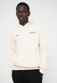 Κρέμ χρώματος φούτερ με μπροστινή τσέπη, ρυθμιζόμενη κουκούλα με κορδόνια και εκτυπωμένο λογότυπο σε σκούρο πράσινο και πορτοκαλί. Σκούρα παντελόνια διακρίνονται από κάτω.