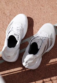 Witte sportschoenen geplaatst op een rode gestructureerde atletiekbaan dicht bij een witte baanslijn, verlicht door natuurlijk zonlicht en werpend schaduwen.