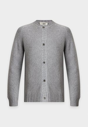Homme debout portant un cardigan gris boutonné sur une chemise blanche, un pantalon foncé à rayures, des chaussures marron et des bijoux en argent.