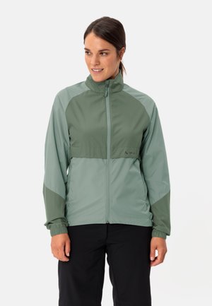 Femme portant une veste zippée bicolore verte avec des poignets élastiques et un pantalon noir, regardant légèrement vers la gauche et souriant.