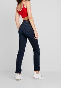 Röd kort axelbandslös topp med vit kant, matchad med mörkblå raka jeans och vita sneakers, sedd bakifrån.