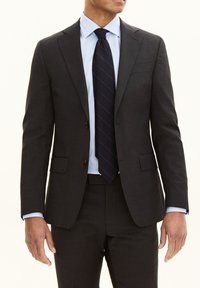 Donkergrijs pak met een op maat gemaakte pasvorm, enkelvoudig knoopjasje, met gelapte revers en twee voorkruiszakken. Gecombineerd met een lichtblauw shirt en een donker gestreepte stropdas.