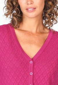 Fuchsia knitted cardigan featuring a textured pattern, V-neckline, and six pink buttons down the front. Soft fabric with visible stitch details.