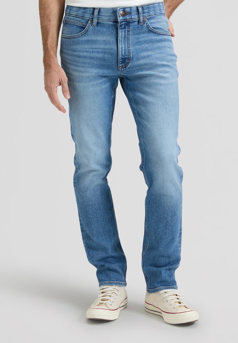 Άνδρας που φοράει στενά τζιν σε ανοιχτό γαλάζιο χρώμα και λευκά αθλητικά παπούτσια, στέκεται μπροστά σε απλό γκρι ανοιχτό φόντο.