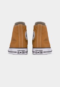 Ζευγάρι καφέ ψηλά αθλητικά παπούτσια Converse All Star, που φαίνονται από πίσω, με λευκές λαστιχένιες σόλες και μαύρο λογότυπο στις φτέρνες.