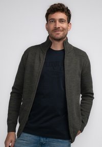 Mörkgrön zip-up cardigan med ribbad krage och axelaccenter, ihop med en marinblå t-shirt. Bärs med ljusblå jeans.