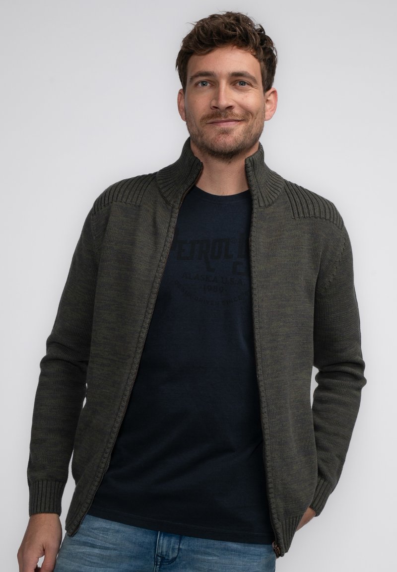 Mörkgrön zip-up cardigan med ribbad krage och axelaccenter, ihop med en marinblå t-shirt. Bärs med ljusblå jeans.