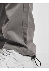 Un pantalon en coton gris présente une coupe ample avec un ourlet retroussé. Associé à des baskets blanches, mettant en valeur une sangle d'ajustement noire au niveau de la cheville.