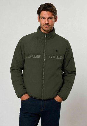 Mężczyzna w ciemnozielonej pikowanej kurtce U.S. Polo Assn., z rękami w kieszeniach, stojący na jednolitym jasnym tle.