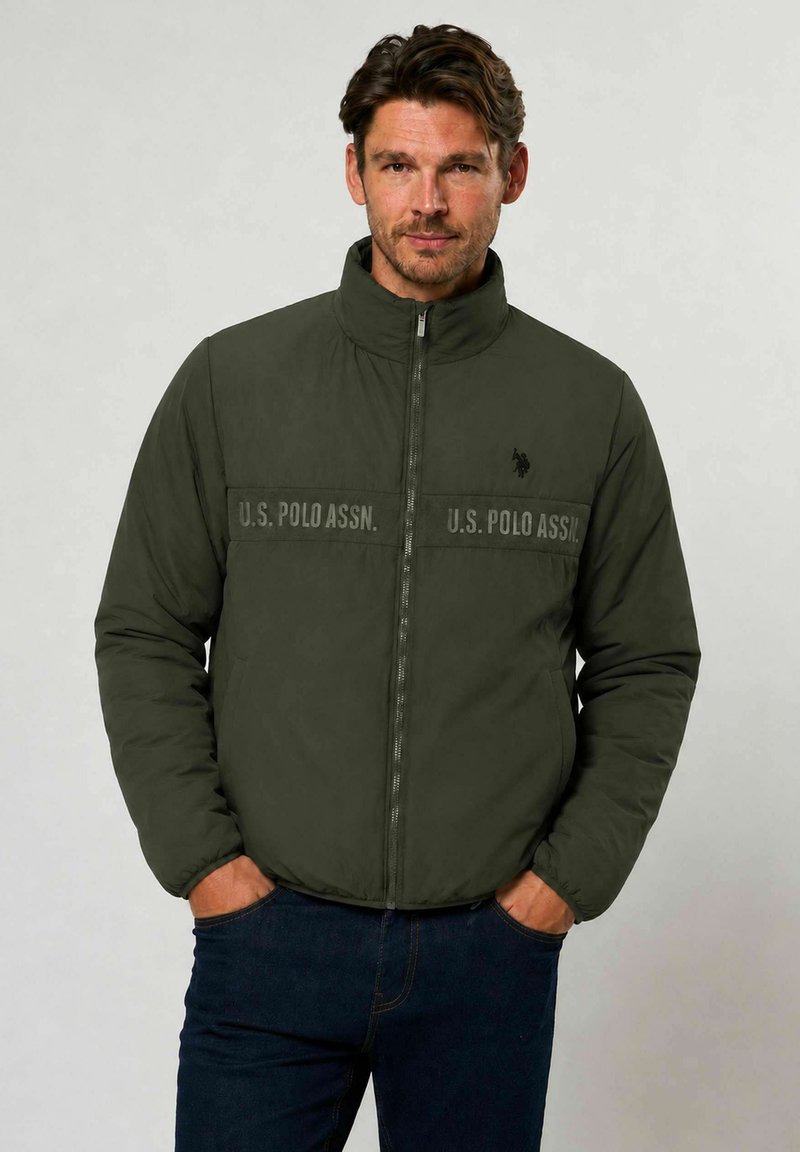 Homme portant une veste matelassée U.S. Polo Assn. vert foncé, les mains dans les poches, debout devant un fond clair uni.