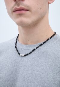 Mustast helmest valmistatud kaelakee hõbedaste aktsentidega, kantud halli särgi peal. Helmeste suurus varieerub, näidates sujuvat tekstuuri.