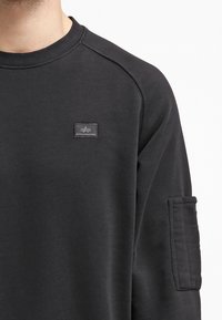 Sudadera negra de algodón con cuello redondo, puños acanalados y un bolsillo lateral con una etiqueta cosida que lleva un detalle de logo.