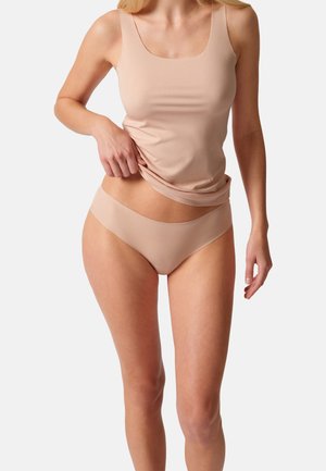 Frau trägt beige nahtlose Unterwäsche und ein Tanktop, zieht den Saum des Tanktops vor einem einfachen weißen Hintergrund hoch.