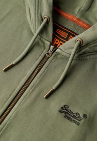 Sudadera con cremallera en color caqui, con cremallera dorada, cordones ajustables y logo bordado en negro en el pecho. Tejido suave y texturizado.