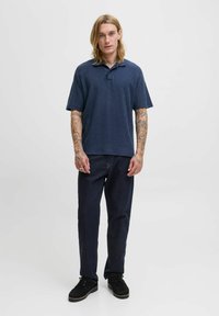 Jūrų laivyno polo marškiniai su tekstūruotu deimanto raštu, derinami su tamsiais džinsais ir juodais raišteliais bateliais. Modelis stovi prieš vienodą foną.
