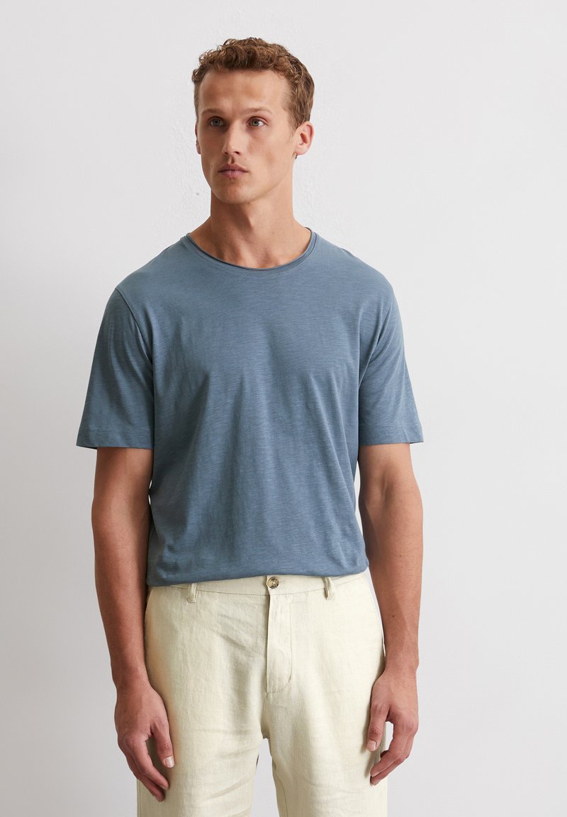 Himmelblaues kurzärmeliges T-Shirt aus weichem, strukturiertem Stoff mit Rundhalsausschnitt, getragen zu beigen Leinenhosen.