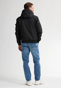 Chaqueta negra con capucha de textura suave, llevada sobre unos jeans de color azul claro. La chaqueta cuenta con puños acanalados y un diseño de corte clásico.