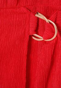 Zbliżenie na czerwony, teksturowany materiał z podwójną klamrą w złotym odcieniu trzymającą marszczony fragment tkaniny.