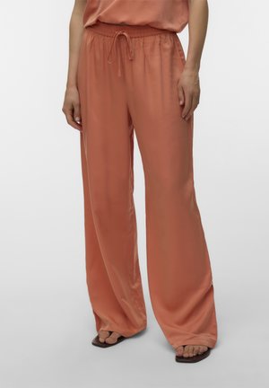 Vrouw draagt losse, wijde broek in terracottakleur met een strikceintuur en sandalen met een vierkante neus op een effen achtergrond.