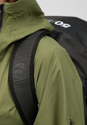 Person iført en olivengrøn jakke med en polstret sort rygsæksrem med en mammutlogo og en sort taske mærket "50".