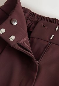 Bordový pás nohavíc s elastickým zadným dielom, strieborný gombík a zips na zapínanie. Obsahuje vertikálnu ryhovanú textúru a kovové akcenty.