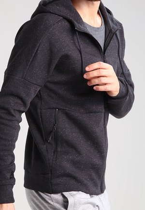 Mand iført en mørkegrå, plettet lynlåshættetrøje med forlomme, der justerer lynlåsen, vist fra midt på torsoen til nakken.