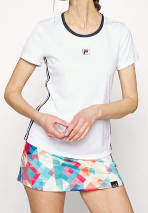 Žena v bílé košili s krátkým rukávem a námořnicky modrým límcem, s vícebarevnou sukní s geometrickým vzorem, se sepjatýma rukama v pase.