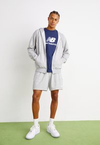Lys grå zip-up hættetrøje over en marineblå t-shirt med hvid branding, parret med grå shorts og hvide sneakers på grønne gulve.