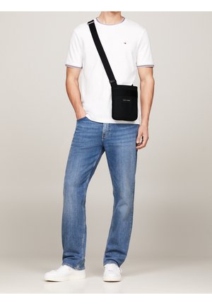 Hombre con camiseta blanca de manga corta, jeans azules, zapatillas blancas y bolsa cruzada negra con correa ajustable, de pie de forma relajada.