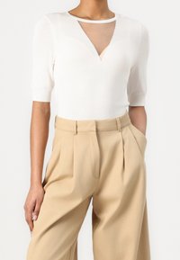 Vit stickad topp med korta ärmar och en V-skärning vid halsen som har en genomskinlig insats. Kombinerad med beige högmidjade vida byxor.