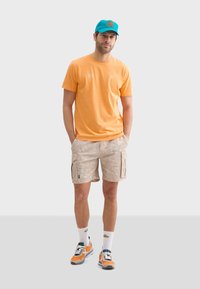 Camiseta de algodón naranja, pantalones cortos de carga beige con patrón de hojas, gorra azul con logo naranja, zapatillas multicolor, calcetines blancos hasta el tobillo.