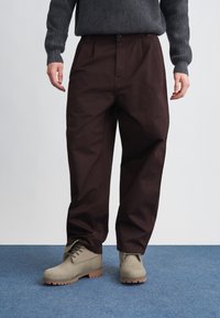Pantalones de algodón marrón con un corte relajado, con pliegues frontales y bolsillos laterales, combinados con botas gris claro sobre una alfombra azul.