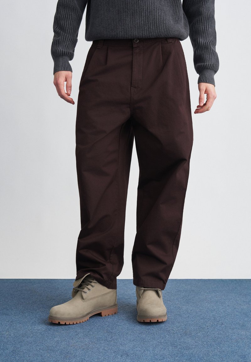 Pantalones de algodón marrón con un corte relajado, con pliegues frontales y bolsillos laterales, combinados con botas gris claro sobre una alfombra azul.