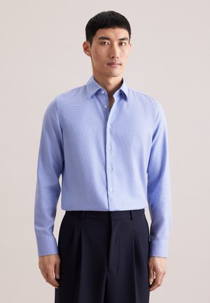 Chemise bleu clair à boutons avec un motif texturé, dotée d'un col italien, de manches longues et de boutons blancs. Portée avec un pantalon foncé.