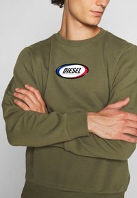 Felpa verde oliva in materiale morbido con polsini e fondo a costine, caratterizzata da un logo ovale "DIESEL" in nero, rosso e blu sul petto.