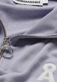 Gros plan sur un tissu violet clair avec une fermeture éclair argentée et un anneau de tirage en métal, montrant le symbole Å brodé en blanc et l’étiquette de la marque « ARMEDANGELS ».