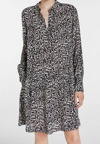 Abito a motivi bianco e nero con maniche lunghe, colletto a button-down e due tasche sul petto. Taglio svasato con vita arricciata. Tessuto leggero.