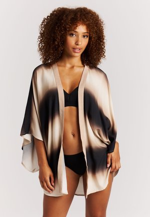 Vrouw met krullend haar die een zwarte bikini en een losse, open kimono-achtige cover-up met een beige-zwart verloop draagt, staand tegen een effen achtergrond.