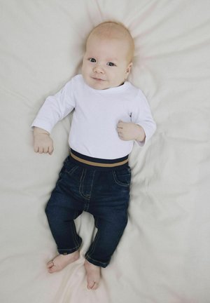 Camisa blanca de manga larga con una cinturilla a rayas en tonos oscuro y claro, combinada con pantalones de mezclilla azul oscuro con costuras amarillas y bolsillos.
