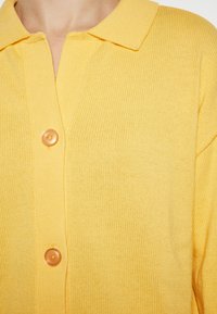 Cardigan giallo lavorato a maglia con colletto e un grande bottone chiaro al centro. Texture morbida, vestibilità comoda.