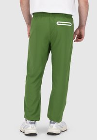 Grüne Sporthose mit glatter Textur, elastischem Bund und einer weißen Reißverschlusstasche auf der Rückseite, kombiniert mit weißen Turnschuhen.