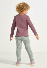 Långärmad lila topp med en vågig nederkant, kombinerad med ljusgröna leggings med ett litet blommönster. Skorna är ljusrosa.