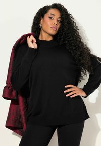 Zwarte gebreide trui met een hoge hals en lange mouwen, gedragen met strakke zwarte leggings en een rood met bloemenpatroon jas die over één schouder is gedrapeerd.