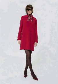 Donna con abito rosso a maniche lunghe, calze nere, scarpe rosse con punta, foulard scozzese al collo, grandi occhiali da sole, in piedi con le gambe incrociate.