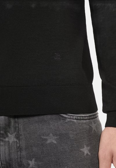 Maglione nero a maglia con orlo a costine, caratterizzato da un piccolo logo impresso. Indossato sopra jeans grigi con motivi a stelle.