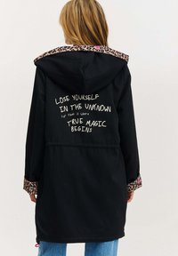 Černý plášť s kapucí a leopardími akcenty na límci a rukávech. Text na zadní straně: "Ztrať se v neznámu, pravá magie začíná."